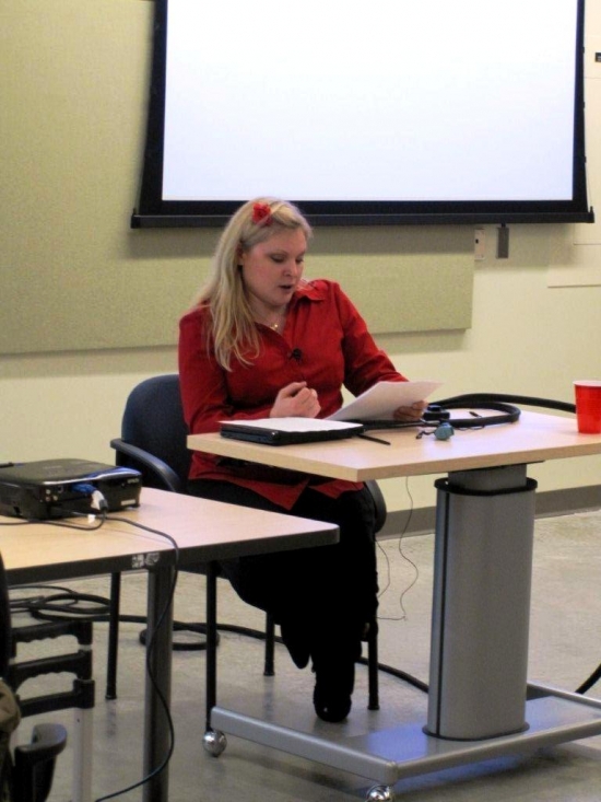 A woman sits and reads from a paper at a desk at the front of the room.