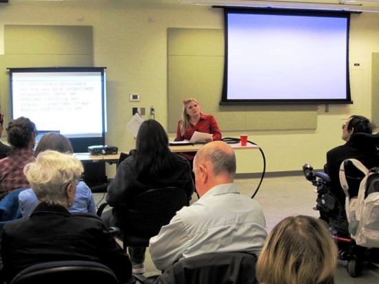 <p>
	A woman sits at a desk while presenting at the front of a large room. Participants sit facing her in the foreground.</p>
