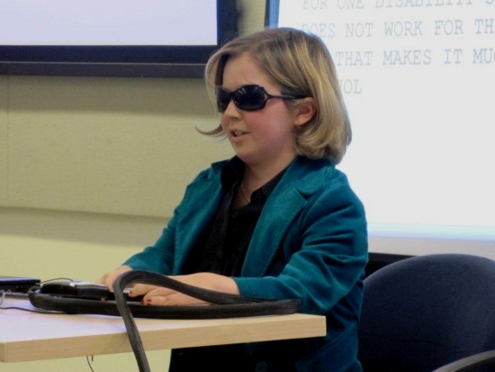 A close-up photo of a woman with sunglasses presenting while sitting at a desk.