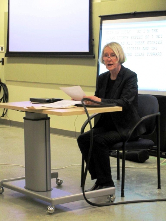 A woman presents while sitting at a desk at the front of a large room.