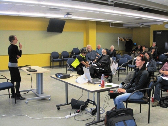 <p>
	A woman stands behind a desk and speaks into a small microphone to seated participants.</p>
