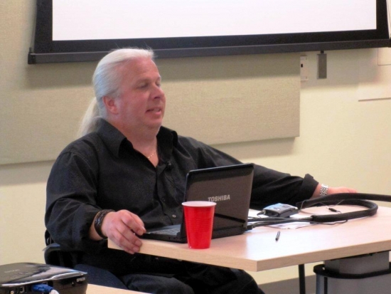 A man sits behind a desk at the front of the room and presents.