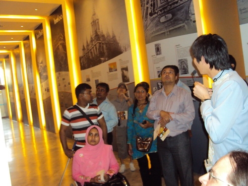Bangladeshi and Nepali team listen to a tour guide in a long hallway filled with photographs and information on Thailand.