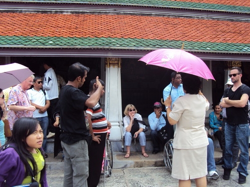 Participants and facilitators sit and stand by the steps of the Grand Palace while listening to a tour guide.