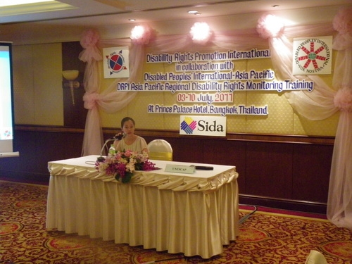 Chona Sabo sits alone at a table and speaks into a microphone. 