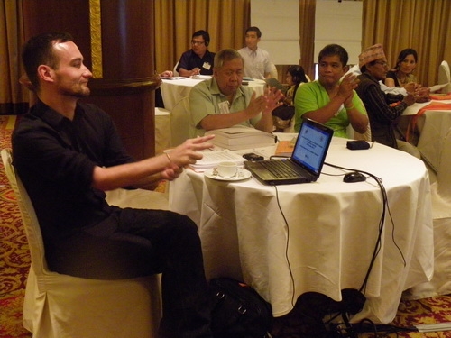 Christopher Lytle, Lauro Purcil and Rannil Sorongon sit at a table with a laptop. Participants sit at tables in the background. 