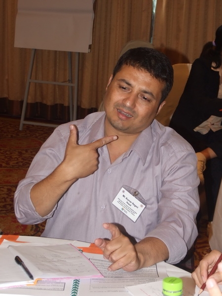 Kumar Regmi points to himself and talks while siting at a table with training materials in front of him. 