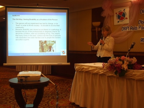 Marcia Rioux, stands beside a slide presentation and speaks into a microphone. 