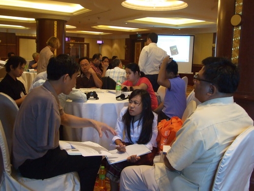 Khamphon, Phoutsady and Lapkeo sit together and have a discussion. There are people and tables in the background. 