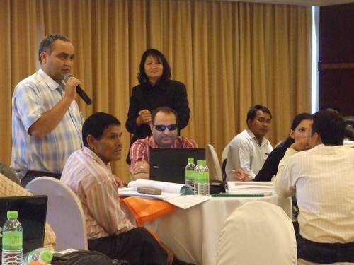 Mr. Harka Bahador stands next to a table with seated participants and speaks into a microphone. 