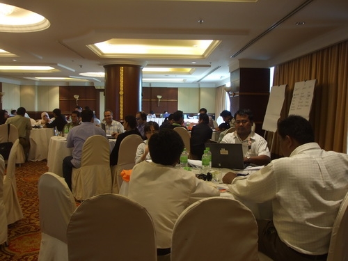 Many participants are seen sitting at tables and working with training materials. 