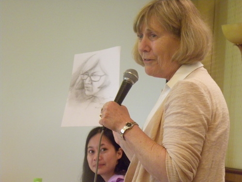 Marcia Rioux holds up a pencil sketch of herself while speaking into a microphone. Chona Sabo looks on.