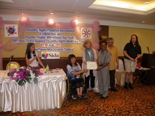 Prum Samoeun smiles and holds his certificate with Saowalak Thonkuay and Marcia Rioux while other facilitators look on. 