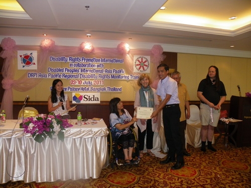 Lapkeo Somchanmavong smiles and holds his certificate with Saowalak Thonkuay and Marcia Rioux while other facilitators look on. 