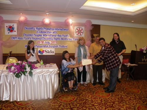 Kumar Regmi smiles and bends with arms outstretched holding his certificate with Saowalak Thonkuay. All the facilitators in the photo are laughing.