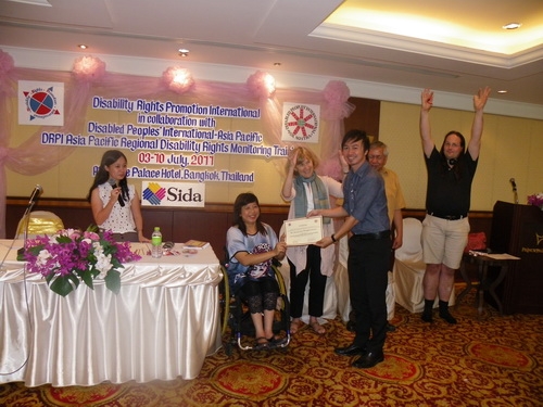 Roong-aran Wongkaewmoon smiles while holding his certificate with Saowalak Thonkuay. Marcia Rioux and Nathan Bond have both their hands raised in the air and smile while other facilitators look on. 