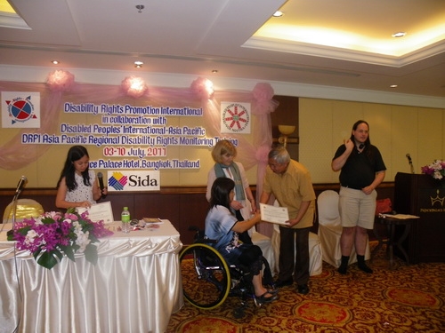 Lauro Purcil holds his certificate and shakes hands with Saowalak Thonkuay while other facilitators look on. 