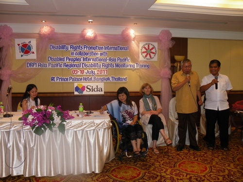 Lauro Purcil stands and speaks into a microphone while other facilitators sit and listen in the background. 