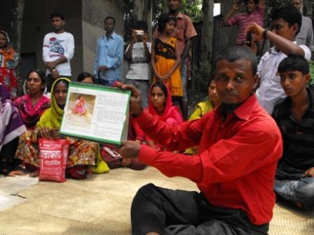 <p>A man from a local DPO seated on the ground holding a book up with a crowd of people seated and standing behind him</p>
