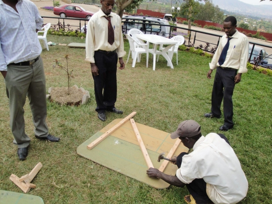 A man kneels to hammer together two boards outside while three other men stand around watching.