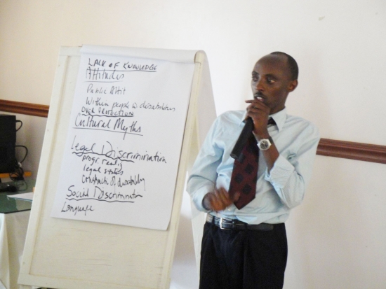 A man speaks into a microphone and stands beside a flip chart showing attitudes people have toward people with disabilities.