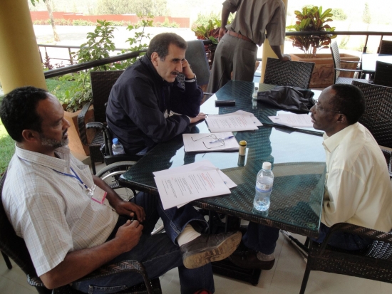 Three men sit at a table covered with training materials on a veranda.