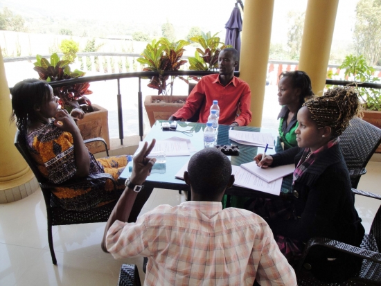 Mr. Ted, Nkatha Murungi, Mercy Mugure, Moses Mayanja and Suzan Kirima sit and talk around a table on a veranda.