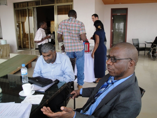 Mr. Robert Mkozho and Jace Nair sit at a table on a veranda. Other participants are in the background standing near a table with coffee carafes.
