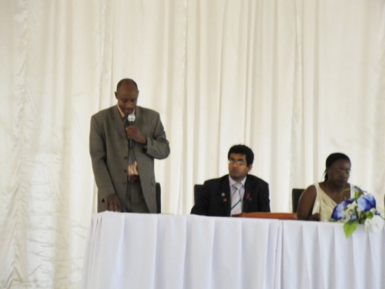 A man in a suit stands and speaks into a microphone with two others seated to the right