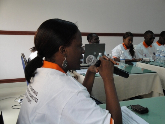 Gloria Namureme speaks into a microphone. Other participants are seated in the background.