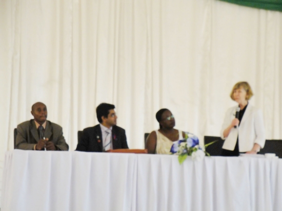 Marcia Rioux stands and speaks into a microphone while 3 others sit to the left