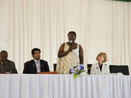 Honourable Minister Jeanne d'Arc Mujawamaliya stands and speaks into a microphone while others sit to her left and right.