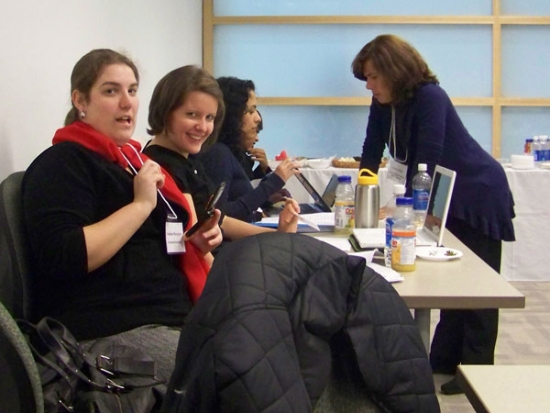 Three women are sitting in a row at a table crowded with food and papers.