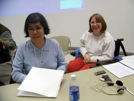 Two participants smile for the photo sit side by side for a photo. On the desk is translation equipment and a bottle of water. 