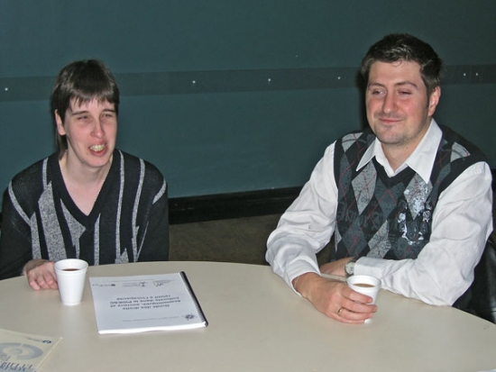 Lyne Robichaud and Jean-Michel Bernier sit at a table and smile while drinking coffee out of styrofoam cups.