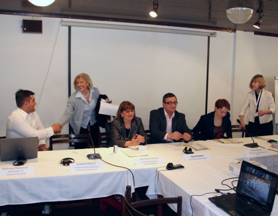 <p>
	Representatives are seated at a long rectangular table adorned with a white tablecloth, microphones and headsets.</p>
