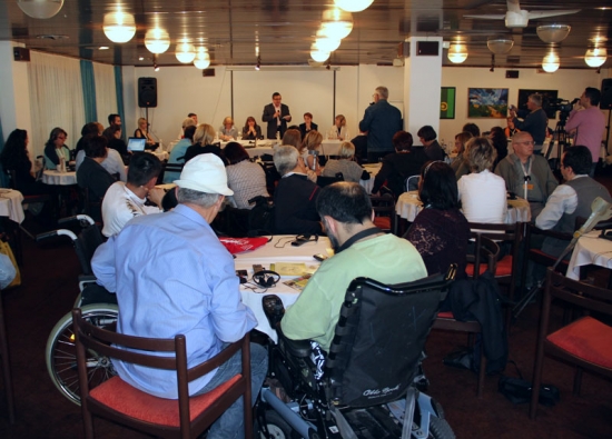 Mr. Mihali stands at the front of the room and speaks into a microphone while training participants sit around tables listening.