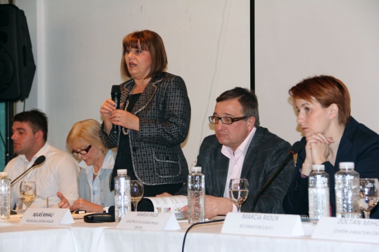 Ms. Petrušić  stands and speaks into a microphone with other organizational representatives seated on either side.