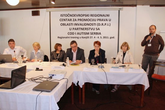 Organizational representatives are seated behind a long table with a white tablecloth. Zoltan Mihok stands to the right, speaking into a microphone.