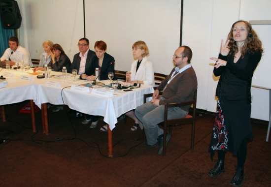 Organizational representatives are seated at a long table. A sign language interpreter is signing to the right of the table.