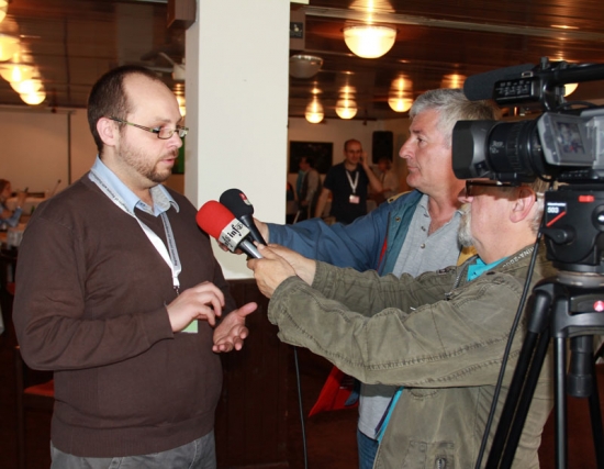 Mr. Zoltan Mihok speaks to two reporters holding microphones. A video camera at the right is filming the interview.