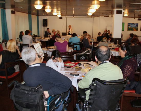 Participants are seated around tables with their backs to the camera. Marcia Rioux is speaking at the front of the room.