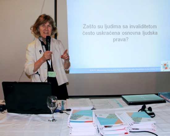 Marcia Rioux stands and speaks into a microphone during a presentation. A powerpoint presentation is projected onto the wall behind Marcia. Training materials are piled on the table in front of her.