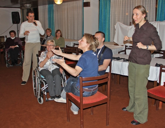 Three participants are seated and smile while a man stands and speaks into a microphone behind them. A sign language interpreter is signing in the foreground.