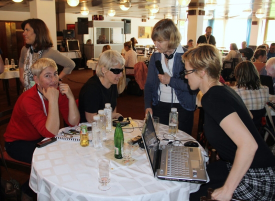 <p>
	Three women sit around a small table with many beverage bottles, headsets and a laptop computer. Marcia Rioux stands beside the table with a microphone.</p>
