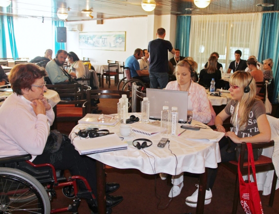 Three women are working at a table covered in water bottles, training material and electronic equipment.