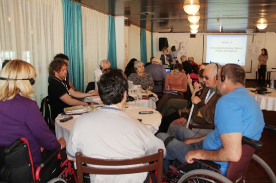 Several men and women are seated around two round tables and while Paula Pinto and Marica Rioux stand at the front of the room. A powerpoint presentation is projected onto the wall behind Paula and Marcia.