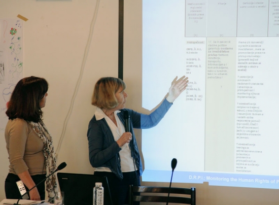 Paula Pinto and Marcia Rioux stand to the side of a slide presentation projected on the wall. Marcia Rioux is gesturing at the slide while speaking into a microphone.