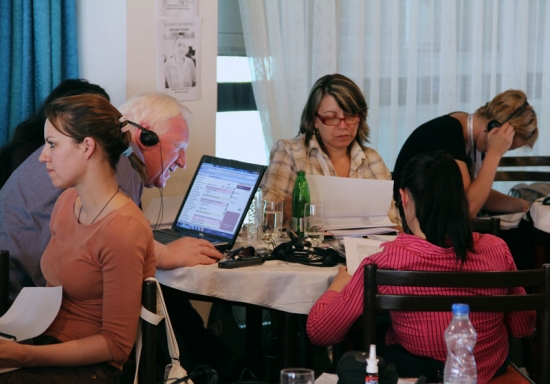 A man and three women are seated around a small round table. The man is works at a laptop computer and the women are looking at papers.