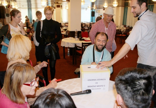 Participants gather around a table with an election box on it. Chris Lytle is placing a piece of paper in the box.